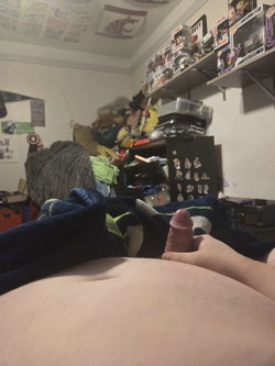 Close-up of a person's lower abdomen and erect penis, hand visible, taken from a low angle in a cluttered bedroom with