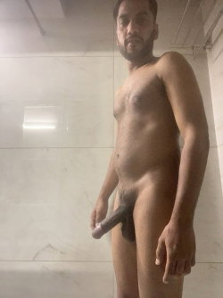 A bearded man with short hair stands nude in a marble-tiled bathroom.
