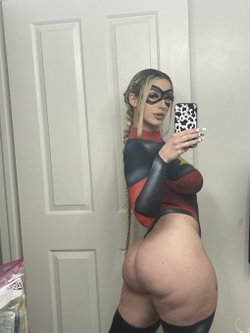 A woman in a tight superhero costume with a mask takes a mirror selfie, showing her bare buttocks and long braided hair.