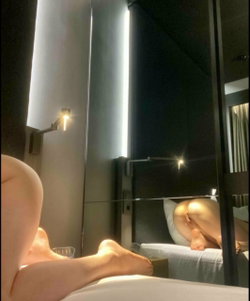 A person with a curvy build lies on a bed in a dimly lit room, partially visible from the legs down, with feet and