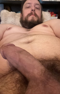 A bearded man lies shirtless on a bed with a white pillow.