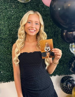 Blonde woman in a black strapless dress smiles while holding a childhood photo, standing in front of a green hedge wall