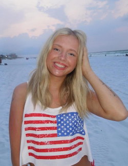 Blonde woman in a white tank top with an American flag print smiles at the camera on a sandy beach at dusk.