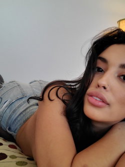 A person with dark hair and a nose ring lies on a bed in a light-colored room.