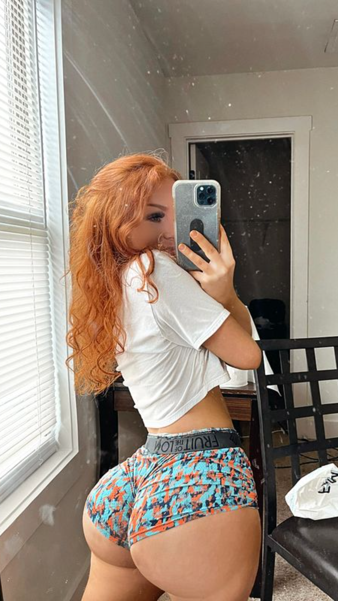 Curvy redhead woman wearing a white crop top and colorful shorts poses for a selfie indoors.
