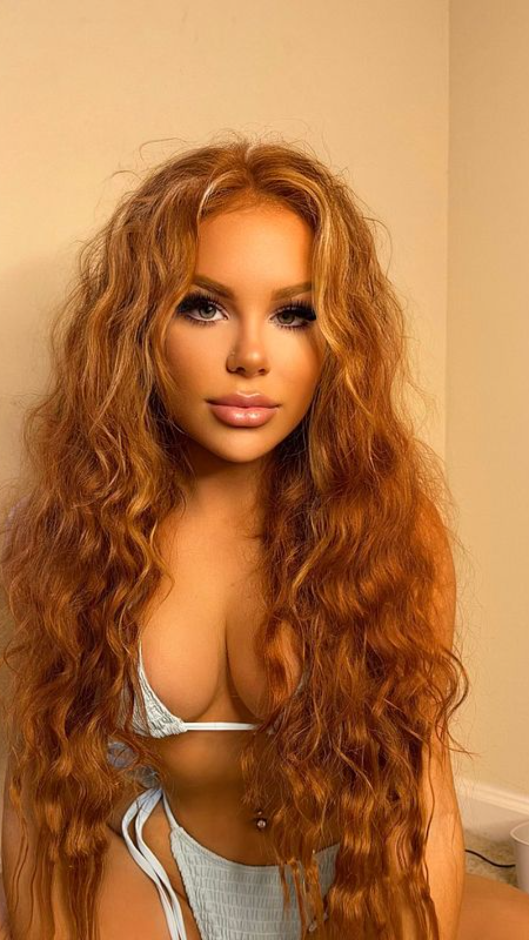 Curvy woman with long wavy auburn hair wearing a light blue bikini indoors.