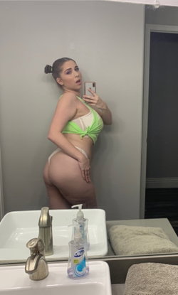 Curvy woman in green and white lingerie takes mirror selfie in bathroom, hand on hip, hair in bun.
