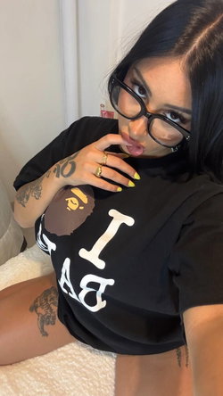 A woman with dark hair and glasses takes a selfie, wearing a black graphic tee, showing tattoos on her arm and thigh