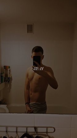 Shirtless man with short hair takes mirror selfie in dimly lit bathroom, wearing striped underwear.