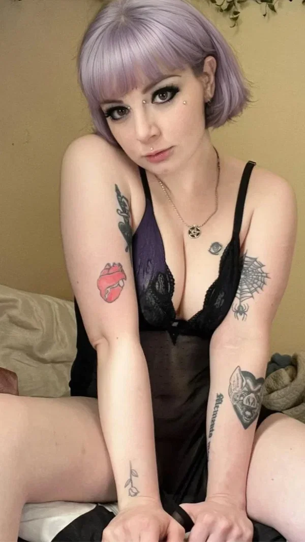 Woman with lavender bob hair and multiple arm tattoos wears a black lace bra and sheer skirt, posing on a bed with