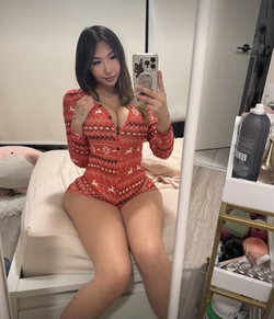 Woman with long dark hair in a red holiday-themed bodysuit sitting on a bed taking a selfie.