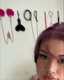 Close-up selfie of a woman with purple hair and nose piercings, standing in front of a wall with various bondage toys