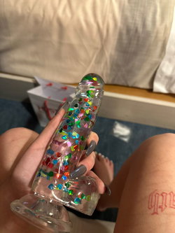 Person holding a clear glitter-filled sex toy, seated on a bed with bare legs and feet visible, one thigh featuring a
