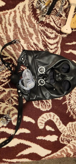 Black leather mask with metal accents, clear toy, and accessories arranged on a patterned rug.