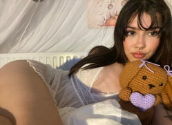 A person with dark hair and a nose ring holds a teddy bear in a white dress.
