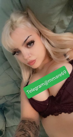 Blonde woman with tattoos takes a selfie in a dark lace bra, lying on a green blanket.