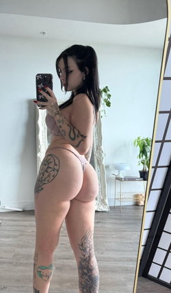 Curvy woman with dark hair and multiple tattoos takes a mirror selfie in a sheer pink top and thong, standing in a