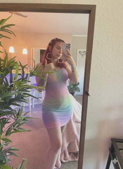 A woman with red hair and tattoos takes a mirror selfie in a rainbow-colored mini dress, posing in a cozy room with