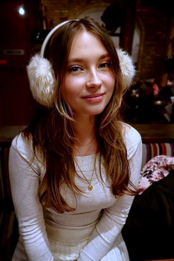 Young woman with long brown hair wearing white earmuffs and a cozy sweater in a cafe setting.
