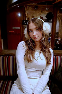Long-haired brunette in a white sweater and earmuffs sits inside a rustic cafe.