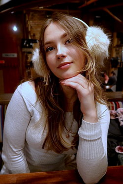 Young woman with long brown hair wearing white earmuffs and an off-shoulder sweater in a wooden lodge setting.