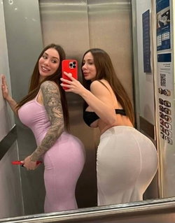 Two women in tight clothing pose for a mirror selfie in an elevator; one wears a pink dress with arm tattoos, the other