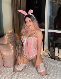 Curvy woman with pink bunny ears, black-pink hair, and lace corset kneels on bed in lit bedroom.