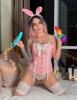 Curvy woman with tattoos in pink bunny corset, garters holds toys on bedroom bed.