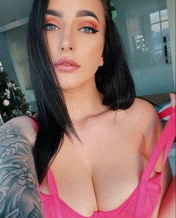 Woman with dark hair and blue eyes wearing a pink top with visible tattoos.