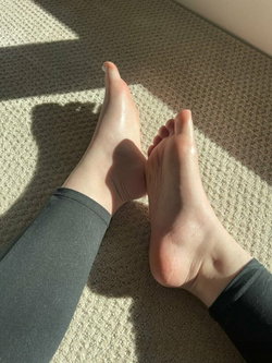 Close-up of bare feet with painted toenails, wearing dark leggings, resting on a textured carpet in sunlight.