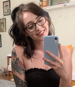A woman with dark hair and glasses takes a mirror selfie in a black strapless top, showing a tattooed arm in a cozy