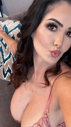 Brunette woman with wavy hair poses in pink lingerie on a couch, making a playful face for the camera.
