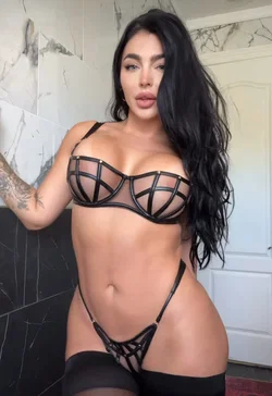 Curvy woman with long dark hair wearing a black strappy lingerie set, posing in a marble-tiled room.
