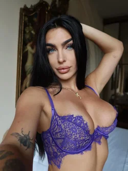 A woman with long black hair and blue eyes wears a purple lace bra, posing indoors with one arm raised behind her head.