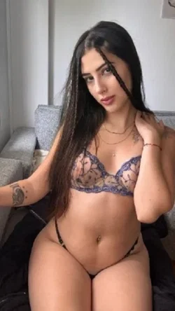 Brunette woman with long dark hair, wearing lace bra and thong, sitting on a gray couch with visible arm tattoos.