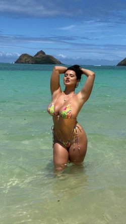 A curvy woman wearing a colorful bikini stands in shallow ocean water with hands behind her head.