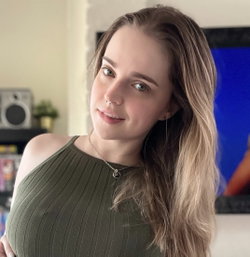 A woman with long wavy hair and a nose ring wears a ribbed olive green halter top, posing indoors near a TV and speaker.