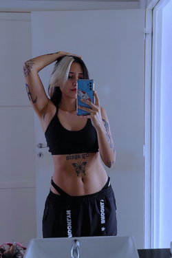 A woman with blonde and dark hair takes a mirror selfie in a black crop top and black shorts, showing tattoos on her