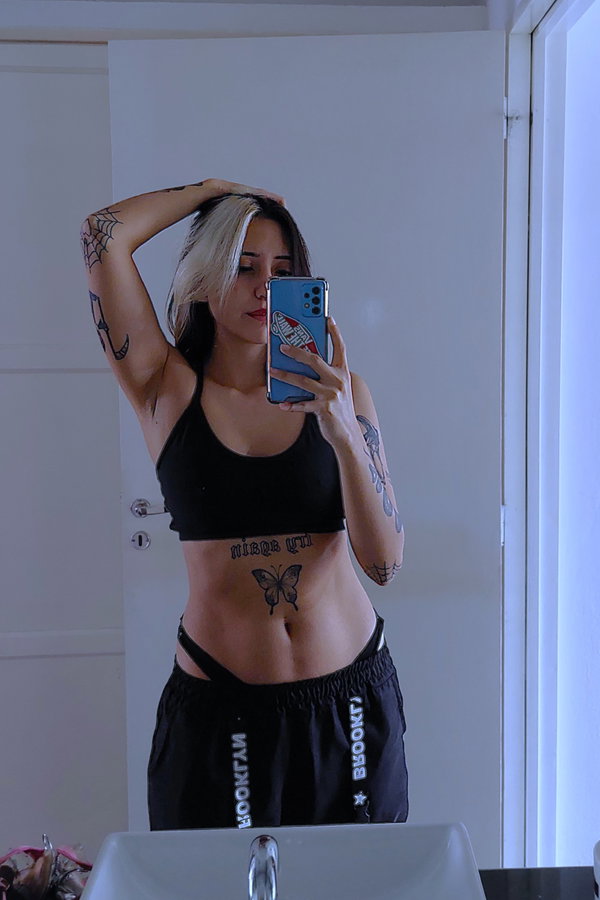 A woman with blonde and dark hair takes a mirror selfie in a black crop top and black shorts, showing multiple tattoos