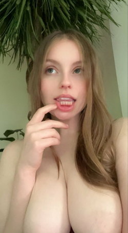 A woman with long light brown hair and fair skin poses topless, touching her lips, in front of green foliage.