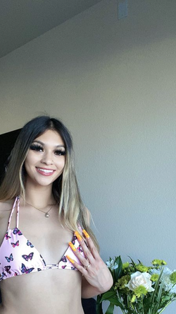 A smiling woman with long ombre hair wears a pink butterfly bikini top indoors near flowers.
