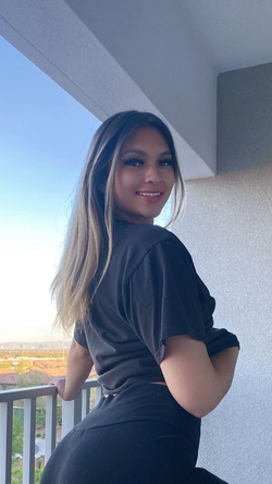 A smiling curvy woman with long highlighted hair poses on a balcony in a black outfit.
