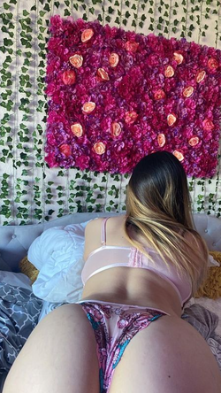 Woman with long highlighted hair in pink lingerie sits on a bed facing a vibrant flower wall.
