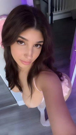 Brunette woman in pink bra and white stockings poses for a selfie in a bedroom.