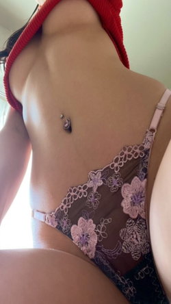 Woman in red hat and floral lace lingerie, showing toned midriff with belly button piercing, indoors near window.