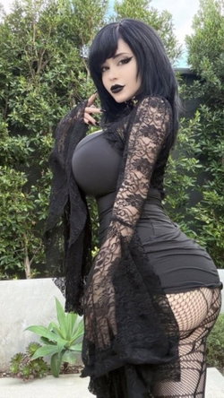Curvy woman with black hair and dark makeup poses in a black lace outfit outdoors.