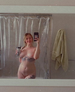 Blonde woman with visible tattoos takes a mirror selfie in a bathroom, holding a drink, bare-chested with lower body