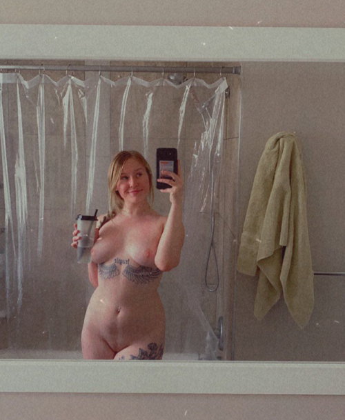 Blonde woman with visible tattoos takes a mirror selfie in a bathroom, holding a drink, bare-chested with lower body