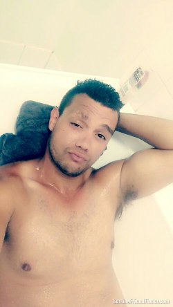 Shirtless man with short dark hair and beard lying in a bathtub, arm resting behind head, water droplets on skin.
