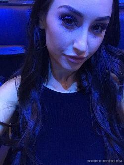 Brunette woman with long dark hair and dramatic eye makeup, wearing a black top, taking a close-up selfie in dim blue